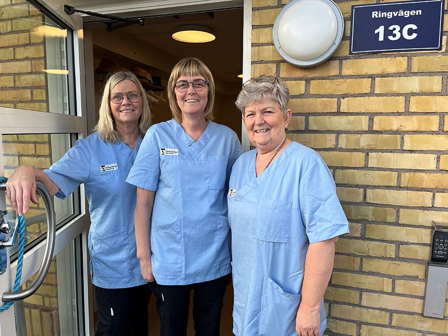 Tre kvinnor i blå arbetströjor står i dörröppningen till en gul tegelbyggnad.