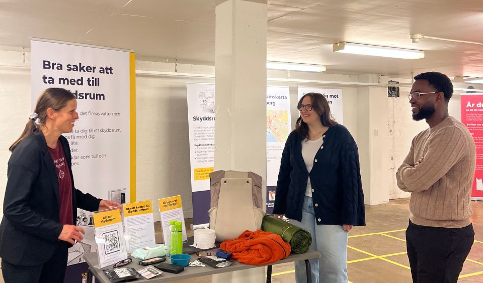 Anja, Linnea och Yassir kring ett bord med saker som kan vara bra att ha med i ett skyddrum, till exempel varma kläder, mat, en radio, våtservetter, radio, toalettpapper med mera.