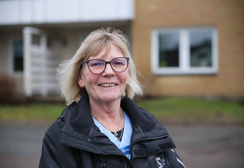 En leende äldre kvinna med blond page och glasögon står framför ett orange tegelhus.