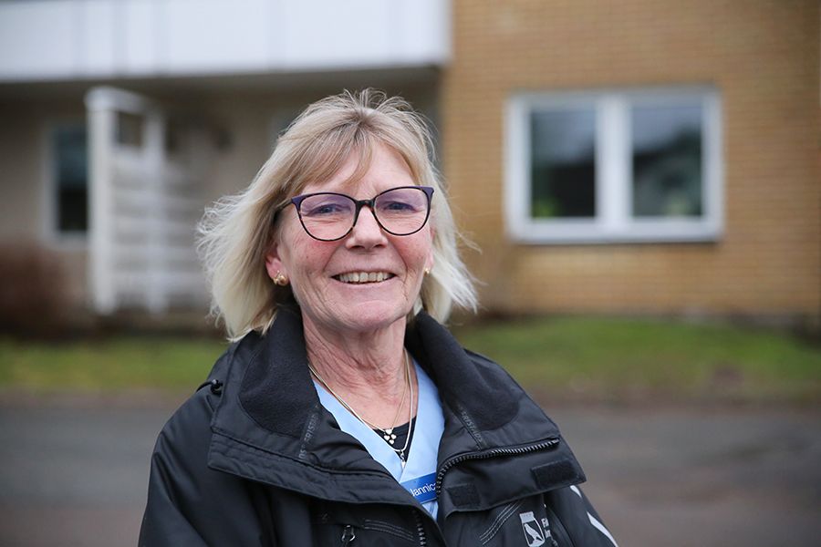 Leende äldre kvinna med blond page och glasögon står framför ett orange tegelhus.