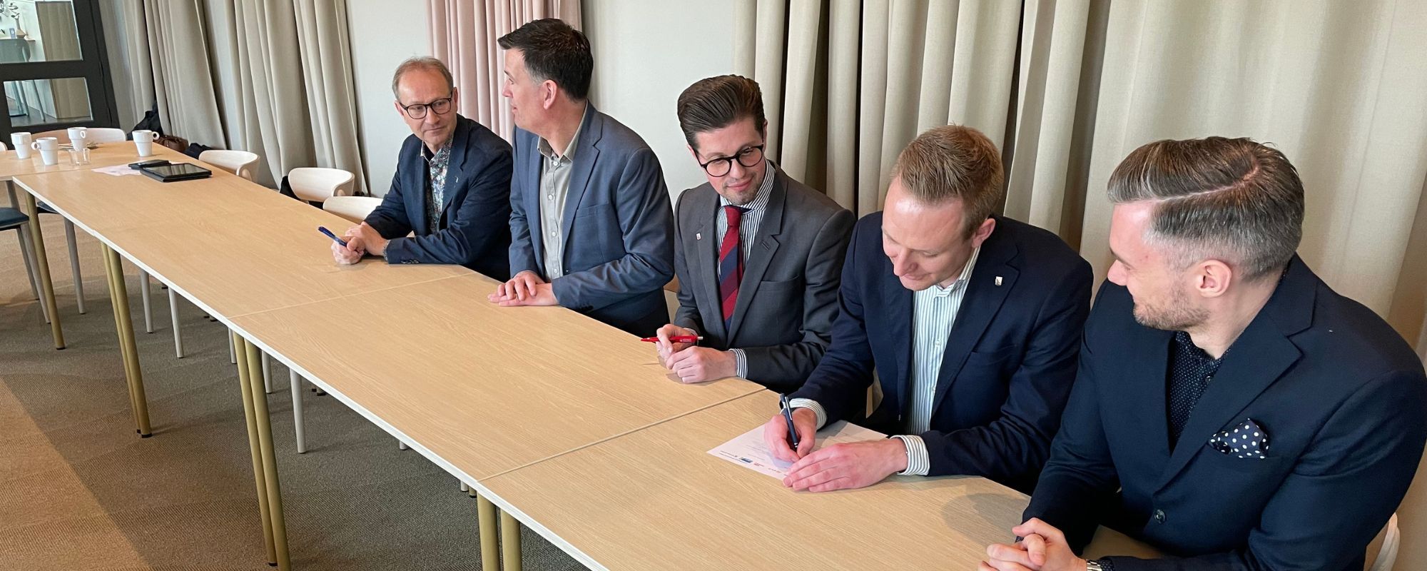 De fem personer som nämns i bildtexten sitter på rad vid ett bord. Alla har mörk kavaj.