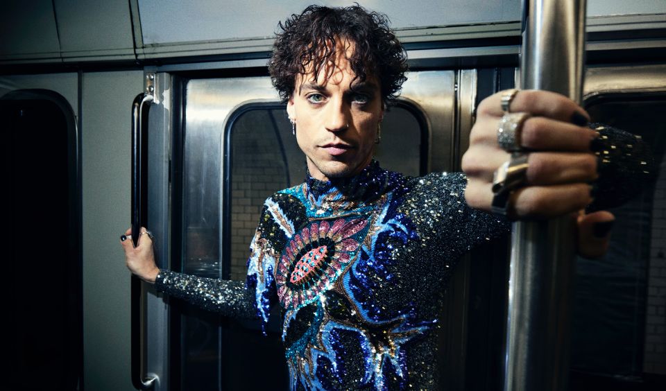 Porträtt i en tunnelbadevagn. Han har en neutral min och tittar in i kameran. Han har en glittrig tröja i blått, svart och lila.