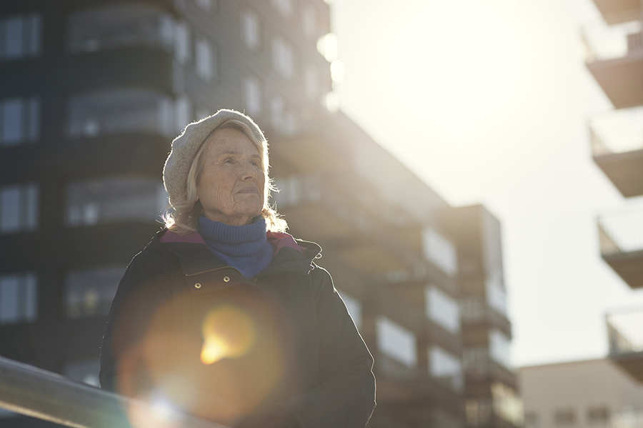 En äldre kvinna står utomhus i ett lägenhetsområde och stirrar ut i luften.