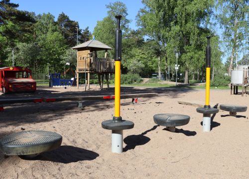 Balansbana i sandlåda, klätterställning och röd träbuss i bakgrunden