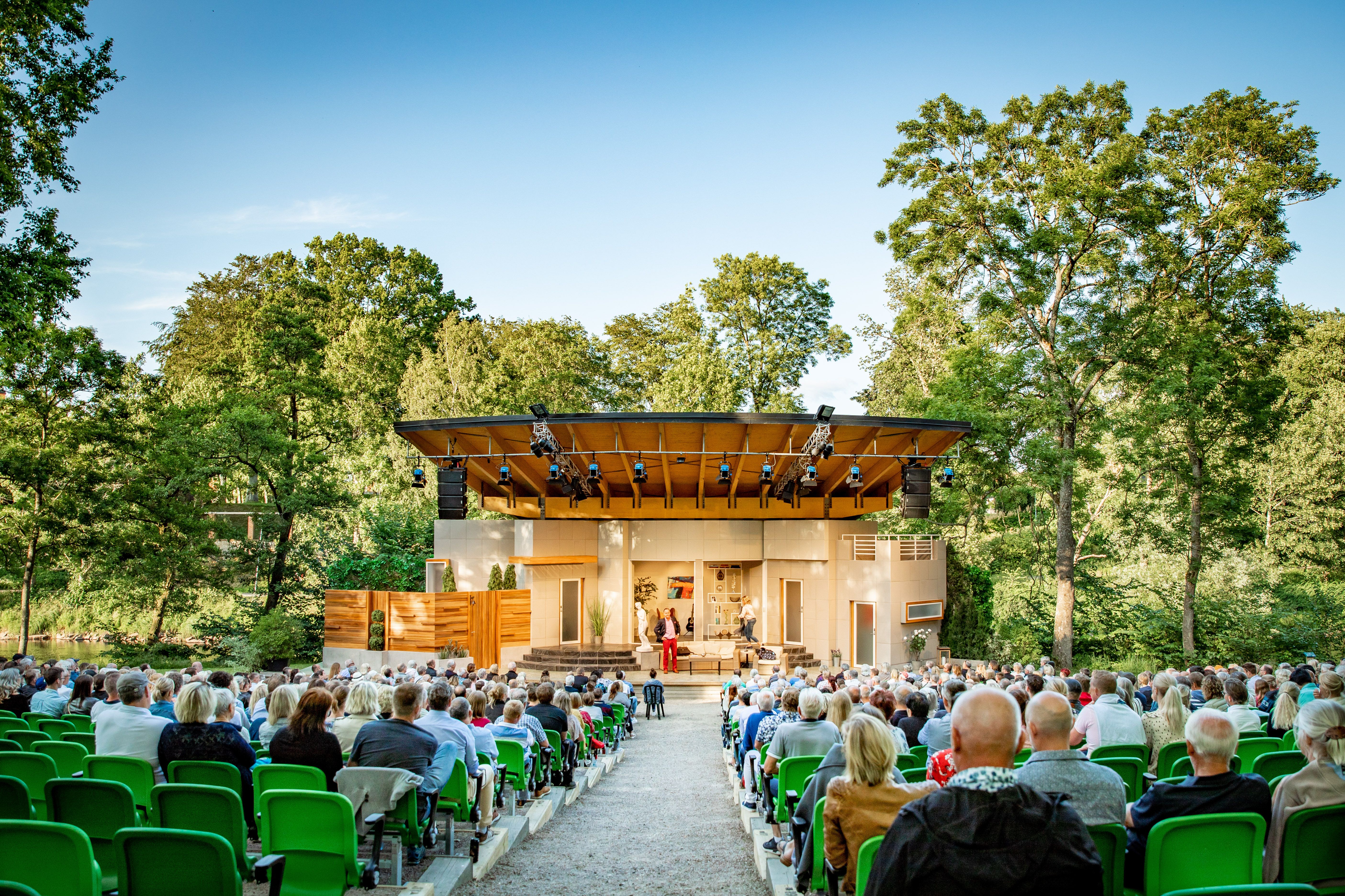 En stor publik sitter framför en utomhusscen. Naturen runt omkring är grönskande.