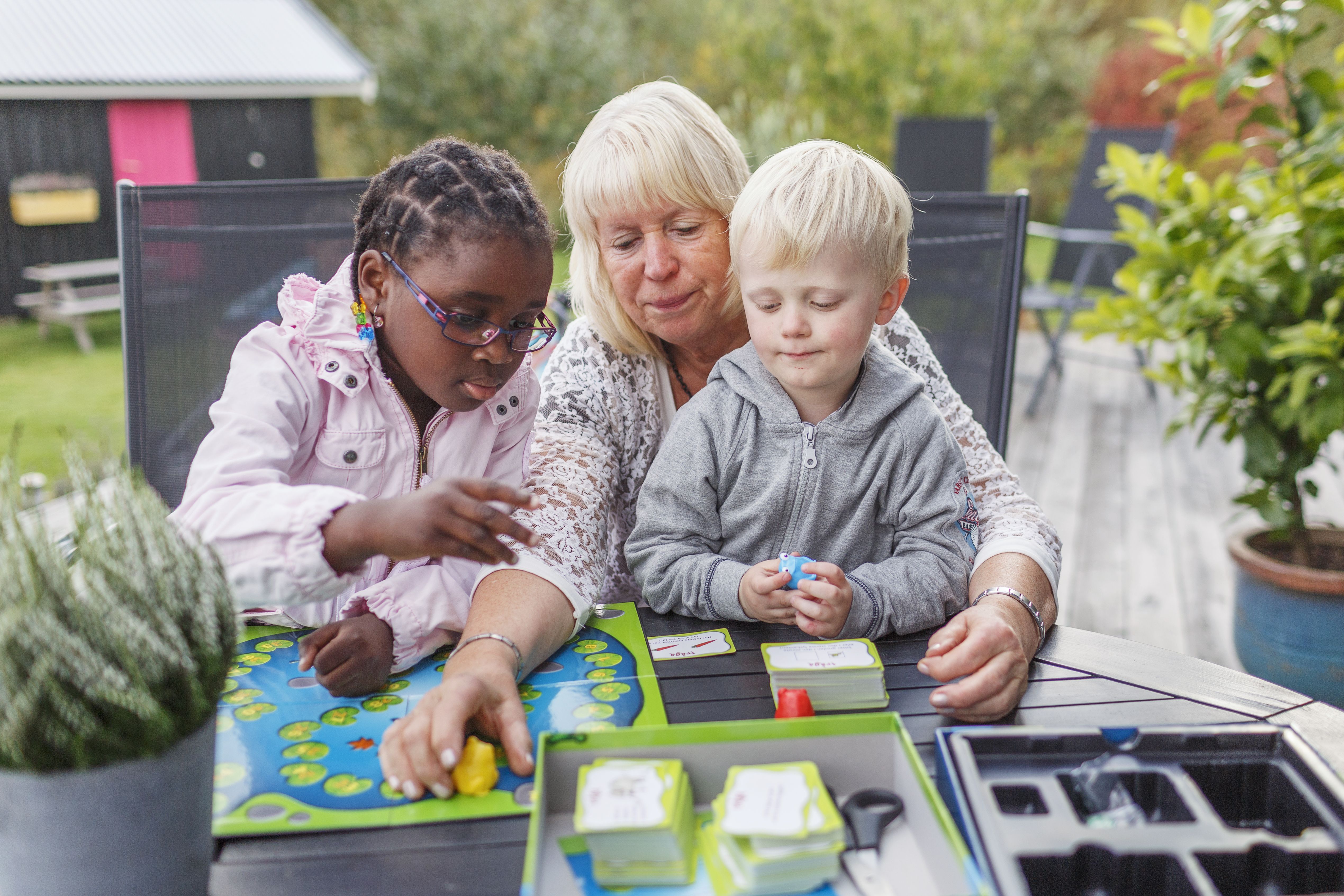 En äldre kvinna spelar spel tillsammans med en mörkhyad flicka och en blond pojke. De sitter utomhus.