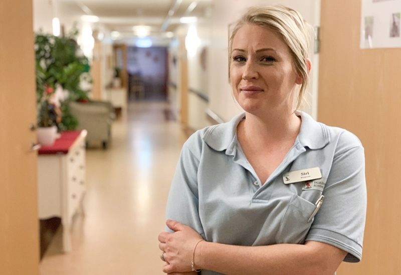 Leende, blond tjej med ljusblå t-shirt står i korridoren på ett särskilt boende för äldre.