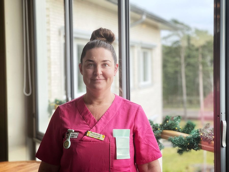 Kvinna med brunt hår uppsatt i knut och rosa vårdkläder står ute på en inglasad veranda.
