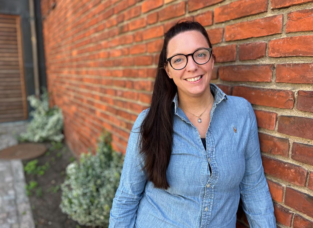 Kvinna i jeansskjorta, glasögon och hår i hästsvans, lutar mot tegelstensvägg.