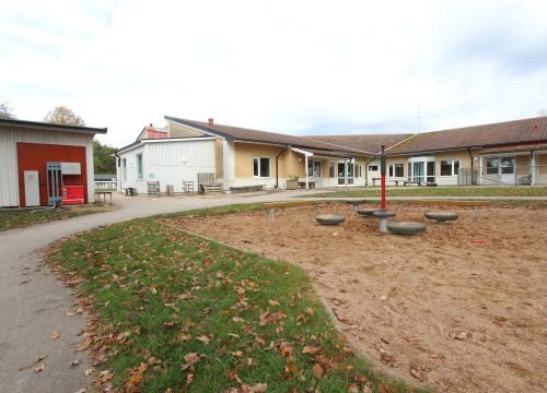 Sandlåda med balansbana, gul låg byggnad i bakgrunden