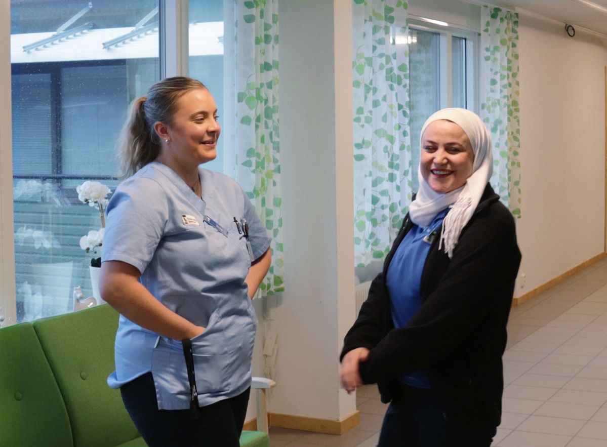 Tova och Farah i korridoren på Solhaga. Tova har ljust hår i hästsvans, Farah har en vit slöja. Båda har hemtjänstens kläder på sig.