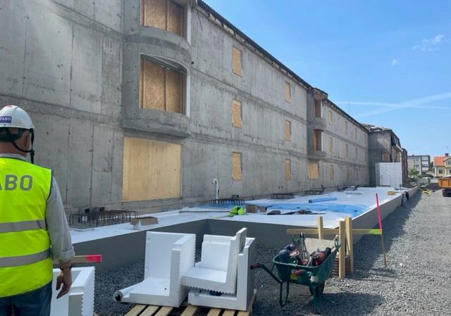 En fastighet med träskivor för fönstren. Till höger om fastigheten syns en stor, avlång form där betongplattan ska gjutas.