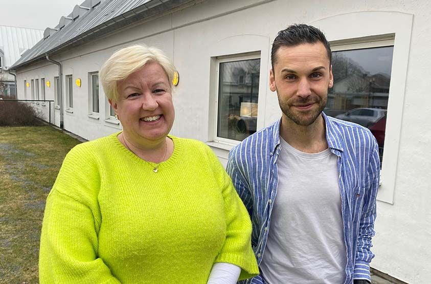 En leende kvinna med kort ljusblont hår och en man med kort brunt hår står framför en vit byggnad.