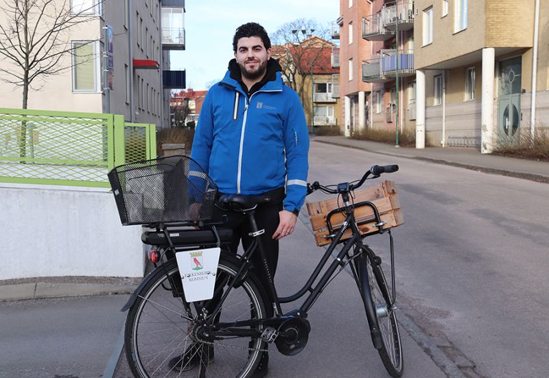 En man med svart hår och blå jacka står ute på en trottoar, bakom en svart cykel.