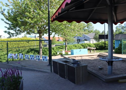 Sandlåda med solskydd, teckningar på staket - buskar och hus i bakgrunden