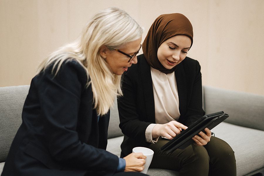 Två kvinnor, en med långt, gråblont hår och en med brun slöja, sitter bredvid varandra i en soffa och tittar på en surfplatta.