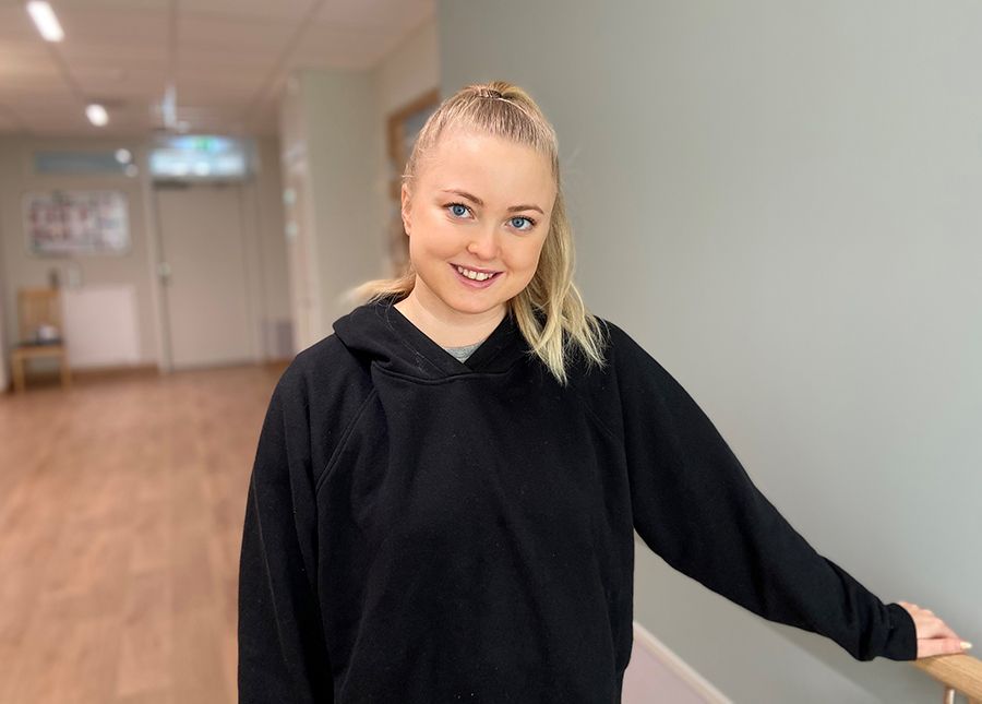 Blond, leende tjej med håret uppsatt i tofs och med svart hoodie står i en korridor.